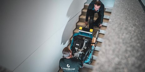In einem hellen Treppenhaus heben zwei Männer eine große Maschine, während sie auf die Stufen achten.