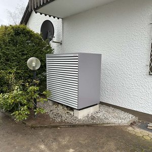 Das moderne Klimagerät mit Lamellen ist in einem Garten platziert, umgeben von Pflanzen und einem beleuchteten Laternenpfahl.