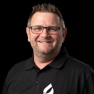 Ein lächelnder Mann mit Brille trägt ein schwarzes Polohemd und steht vor einem dunklen Hintergrund.