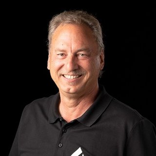 Ein lächelnder Mann mit kurzen, grauen Haaren trägt ein schwarzes Polohemd und steht vor einem dunklen Hintergrund.