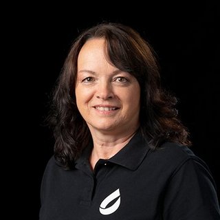 Eine Frau mit lockigem, braunem Haar lächelt freundlich in die Kamera, während sie ein schwarzes Polohemd trägt.