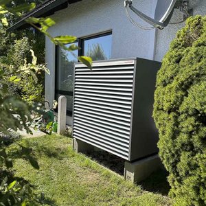 Das Bild zeigt ein kompaktes Klimagerät mit Lamellen, platziert an der Wand eines Hauses, umgeben von Sträuchern.