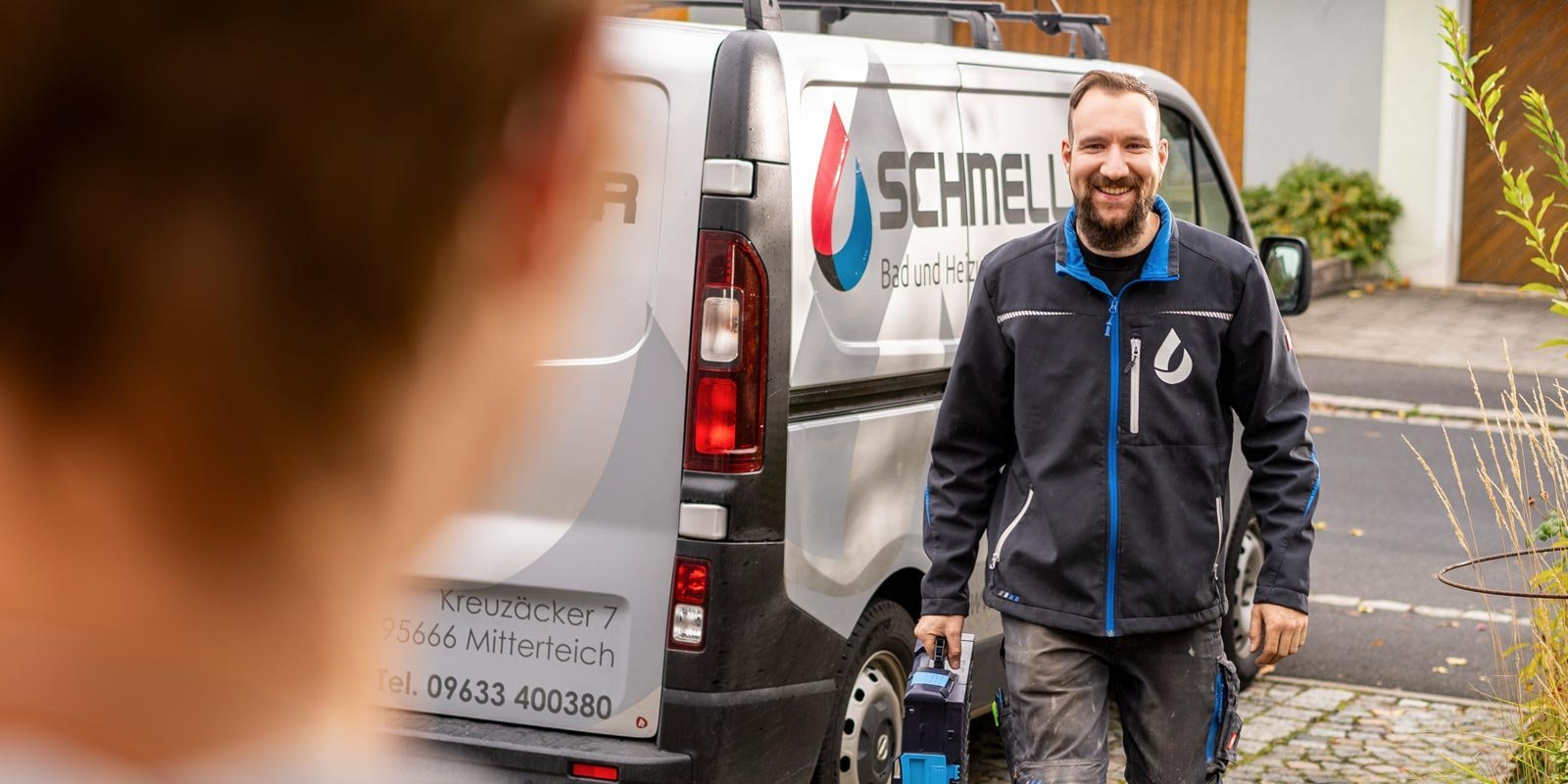 Ein Handwerker mit einem Werkzeug in der Hand steht vor einem Firmenfahrzeug und strahlt Zuversicht und Freundlichkeit aus.