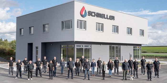 Eine Gruppe von Mitarbeitern steht vor einem modernen Gebäude mit dem Logo 'Schmeller', umgeben von einer grünen Landschaft.