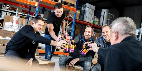  Fünf Männer stoßen mit Flaschen in einer Lagerhalle an, während sie lächeln und eine entspannte Atmosphäre genießen. 