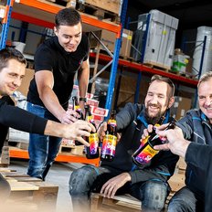  Fünf Männer stoßen mit Flaschen in einer Lagerhalle an, während sie lächeln und eine entspannte Atmosphäre genießen. 