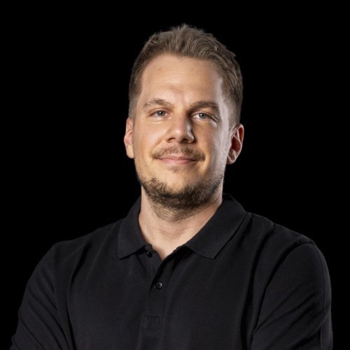 Ein Mann mit kurzem, blondem Haar lächelt selbstbewusst in die Kamera, während er seine Arme vor der Brust verschränkt.