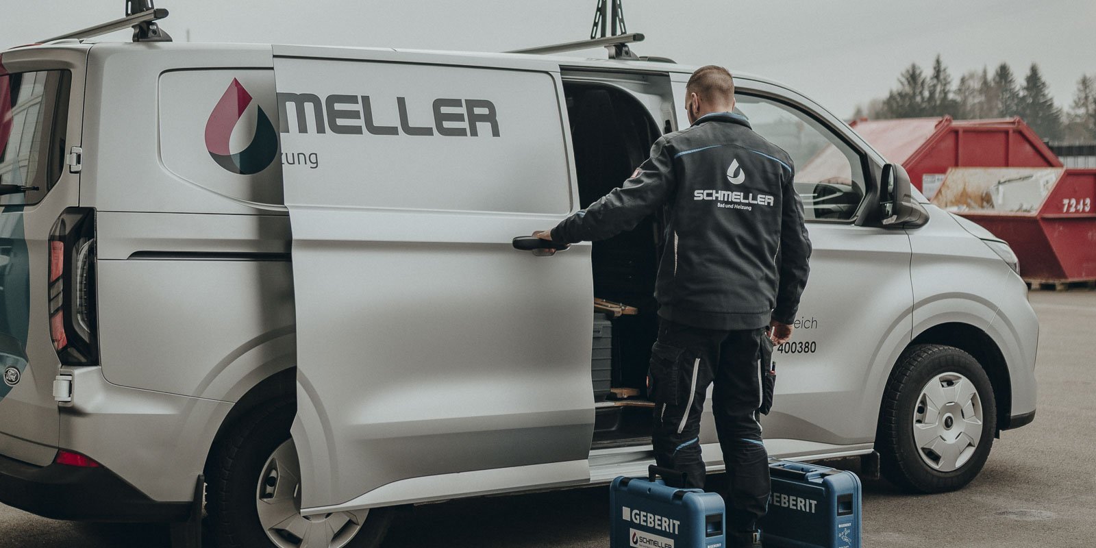 Ein Handwerker in Arbeitskleidung lädt Werkzeuge aus einem Lieferwagen mit dem Logo einer Sanitärfirma aus.