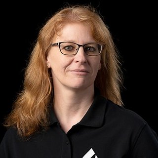 Eine Frau mit langen, lockigen, blonden Haaren und Brille, die in einem schwarzen Poloshirt vor einem dunklen Hintergrund steht.