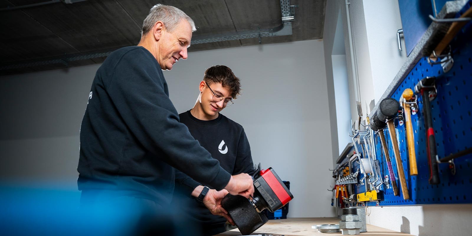In einer modernen Werkstatt zeigen ein älterer und ein jüngerer Mann Freude beim Arbeiten mit Werkzeugen und Maschinen.