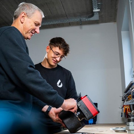 In einer modernen Werkstatt zeigen ein älterer und ein jüngerer Mann Freude beim Arbeiten mit Werkzeugen und Maschinen.