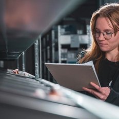In einem modernen Lager schaut eine Frau konzentriert auf ein Dokument, während sie zwischen Regalen steht.