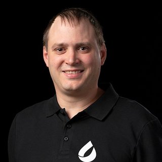 Ein lächelnder Mann mit kurzen Haaren trägt ein schwarzes Polohemd mit einem weißen Tropfen-Logo auf der linken Brust.