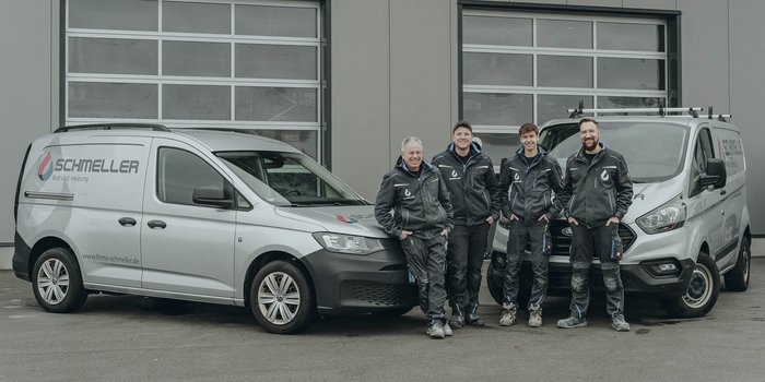 Vier Handwerker in Arbeitskleidung stehen lächelnd vor zwei Firmenfahrzeugen mit dem Logo von Schmeller.