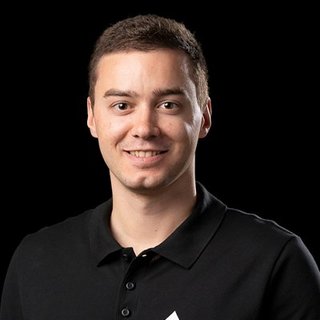 Ein junger Mann mit kurzem, braunem Haar lächelt freundlich in die Kamera, während er ein schwarzes Polohemd trägt.