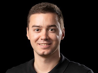 Ein junger Mann mit kurzem, braunem Haar lächelt freundlich in die Kamera, während er ein schwarzes Polohemd trägt.