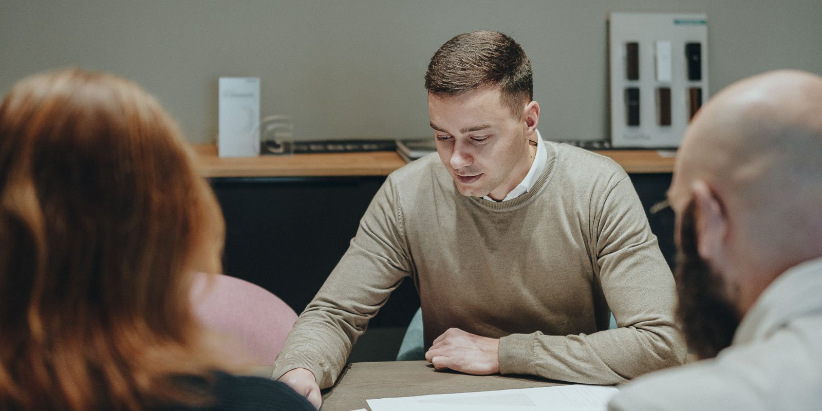 Ein junger Mann in einem beigen Pullover konzentriert sich auf Dokumente auf einem Tisch, während er mit anderen spricht.