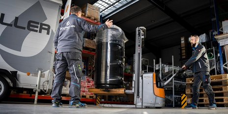 Ein Mitarbeiter steuert einen Gabelstapler, um einen schweren, schwarzen Behälter zu bewegen, während ein Kollege ihn anweist.