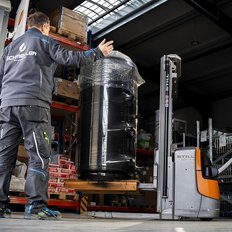 Ein Mitarbeiter steuert einen Gabelstapler, um einen schweren, schwarzen Behälter zu bewegen, während ein Kollege ihn anweist.