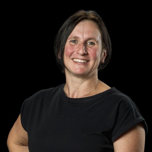 Eine lächelnde Frau mit kurzen, dunklen Haaren trägt ein schwarzes T-Shirt und steht vor einem schwarzen Hintergrund.