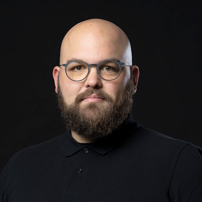 Ein Mann mit Glatze und Bart trägt eine schwarze Poloshirt und eine Brille, während er ernst in die Kamera schaut.