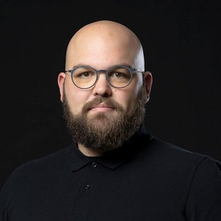 Ein Mann mit Glatze und Bart trägt eine schwarze Poloshirt und eine Brille, während er ernst in die Kamera schaut.