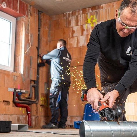 In einem Baustellenraum schneidet ein Mann konzentriert ein Rohr, während ein anderer Arbeiter an einer Wand arbeitet.