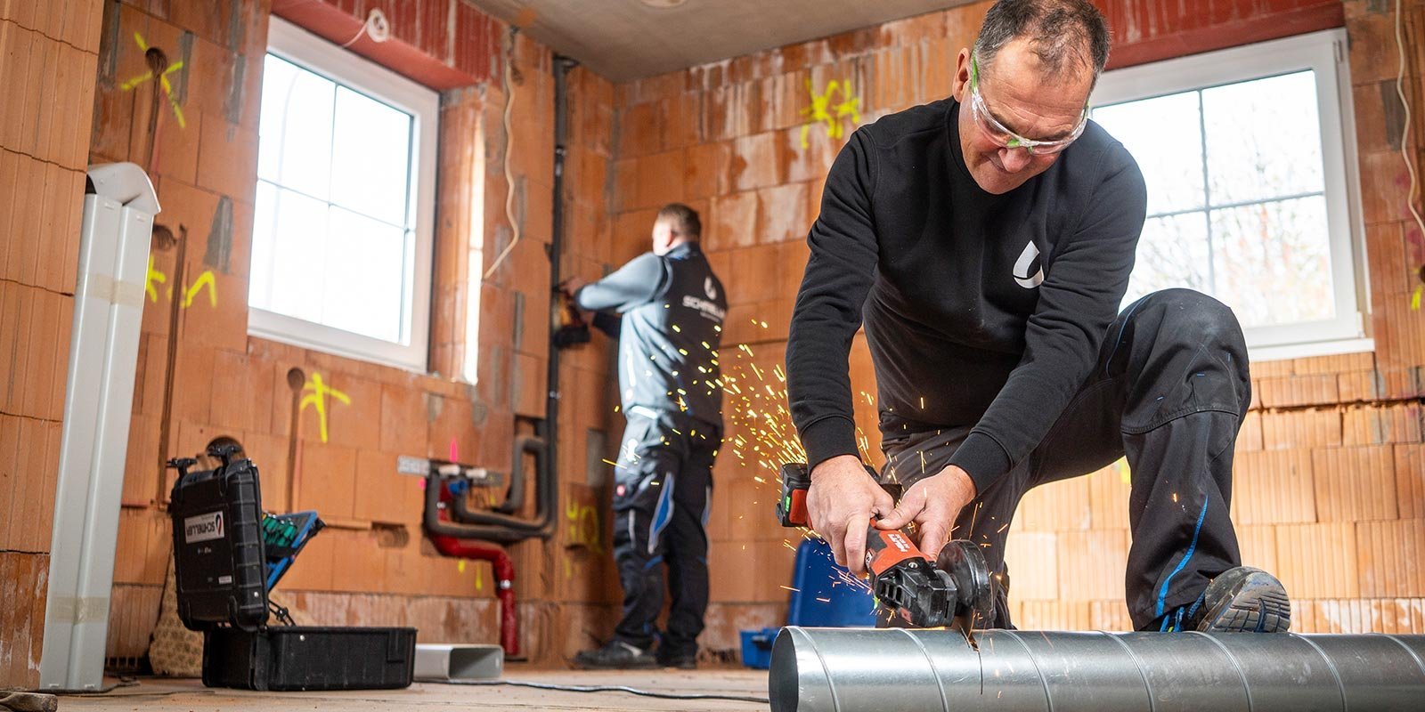 In einem Baustellenraum schneidet ein Mann konzentriert ein Rohr, während ein anderer Arbeiter an einer Wand arbeitet.